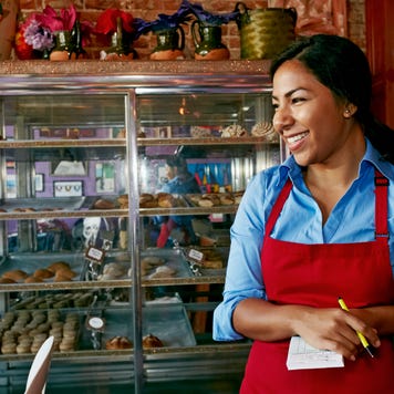 A bakery owner, wearing a red apron, smiles as she stands at the counter.