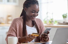 Online Payments. Cheerful black lady using smartphone and credit card in kitchen at home, smiling african woman sitting at table with laptop, paying bills in internet or transferring money via app