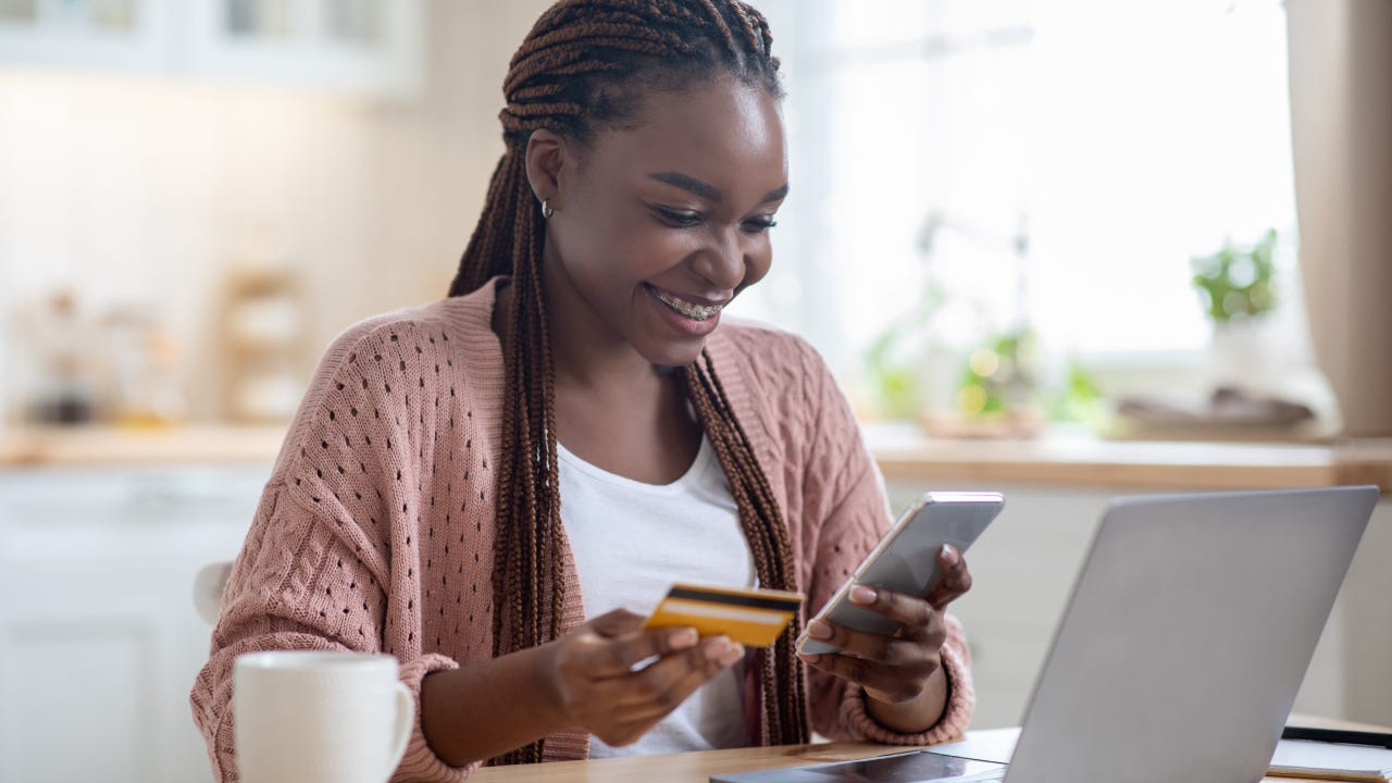 Online Payments. Cheerful black lady using smartphone and credit card in kitchen at home, smiling african woman sitting at table with laptop, paying bills in internet or transferring money via app