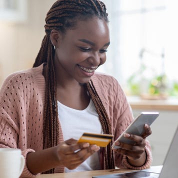 Online Payments. Cheerful black lady using smartphone and credit card in kitchen at home, smiling african woman sitting at table with laptop, paying bills in internet or transferring money via app