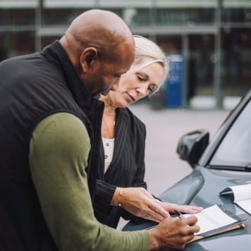 Saleswoman assisting male customer while doing paperwork standing near car