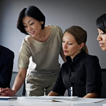 A businesswoman leans over a table, looking over papers with her diverse staff