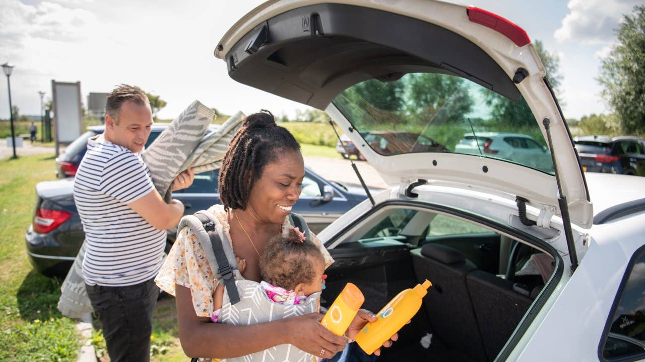 A mother is carrying her baby in a carrier and is holding two types of sun tan lotion. her husband is holding the picnic blanket.