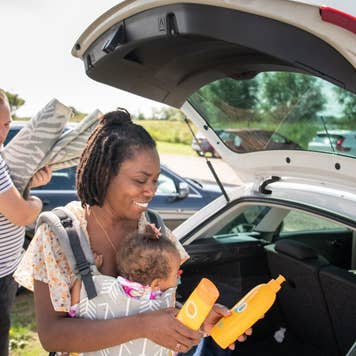 A mother is carrying her baby in a carrier and is holding two types of sun tan lotion. her husband is holding the picnic blanket.