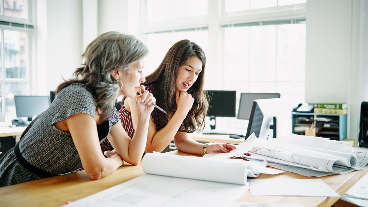 Two women lean on a table reviewing blueprints.