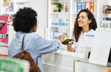 Female customer doing nfc payment with her credit card at pharmacy. Smiling cashier holding credit card reader at drug store checkout counter.