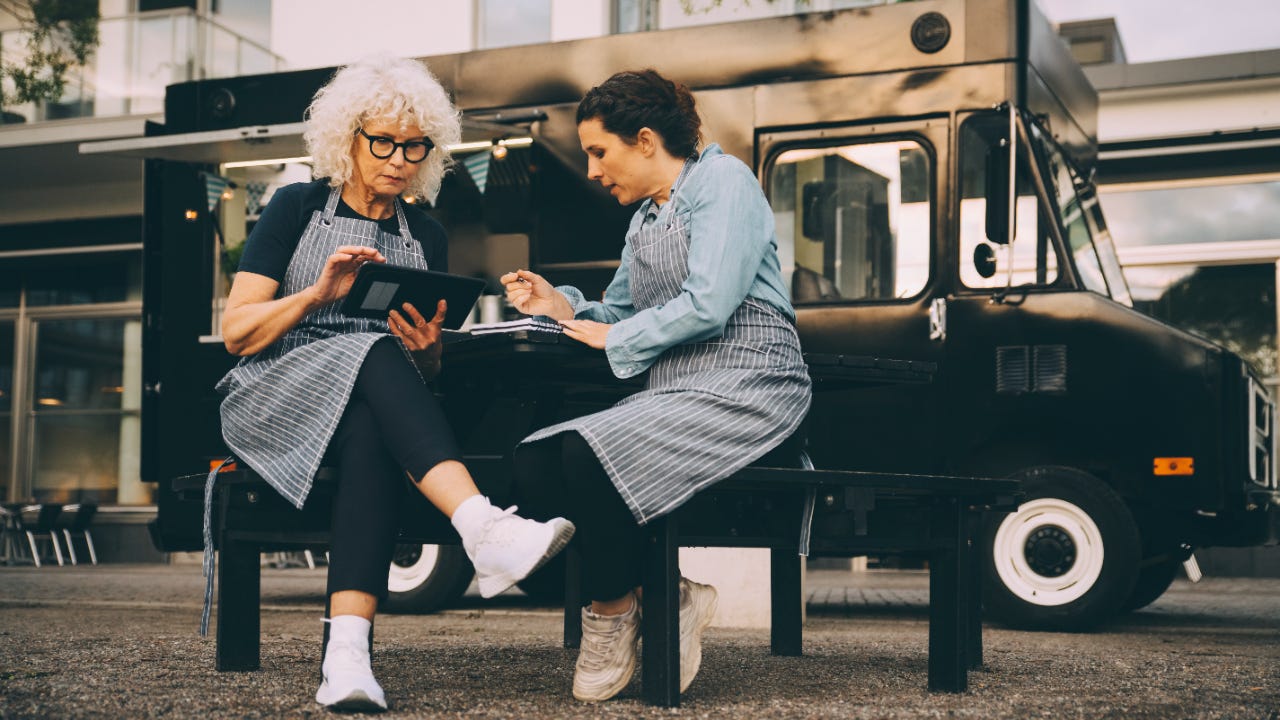 Two women review a table and notes while sitting outside a food truck.