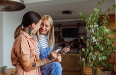 Two women smile at one another. One sits on a table holding a phone, while the other stands holding a credit card.