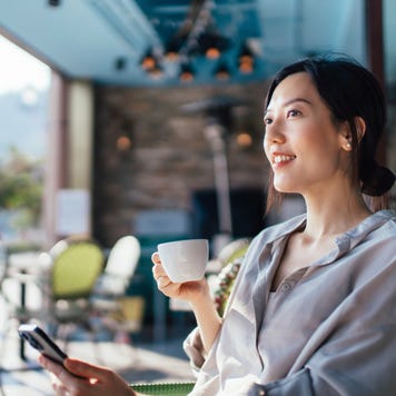Cheerful young Asian woman using smartphone while sitting at a sidewalk cafe.