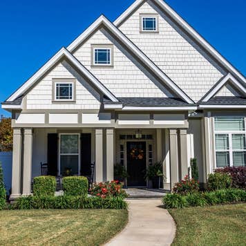 The front view of a cottage craftsman style white house with a triple pitched roof with a sidewalk, landscaping and curb appeal