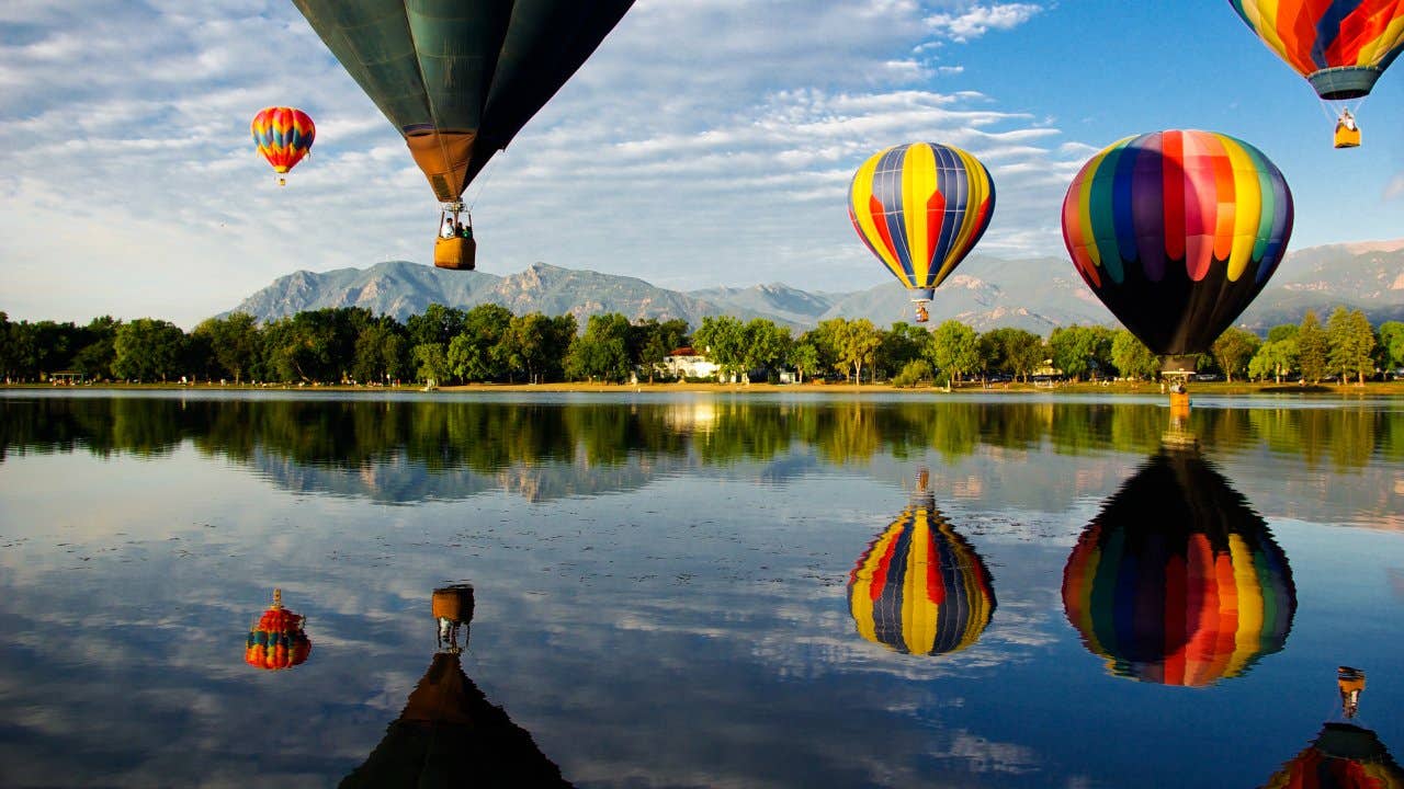 Colorful hot air balloons glide over Prospect Lake in Colorado Springs, CO, at the Colorado Balloon Classic; Cheyenne Mountain is in the background.