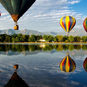 Colorful hot air balloons glide over Prospect Lake in Colorado Springs, CO, at the Colorado Balloon Classic; Cheyenne Mountain is in the background.