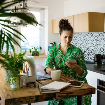 While looking at her cellphone, a young woman sits at her kitchen table with a notebook, tablet, and laptop.