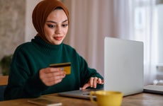 Young woman wearing hijab smiling and shopping online, looking at credit card and keying in card details on laptop at desk