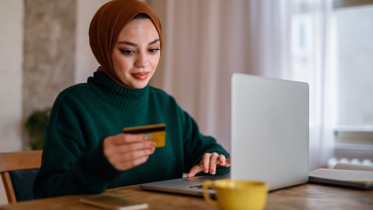 Young woman wearing hijab smiling and shopping online, looking at credit card and keying in card details on laptop at desk