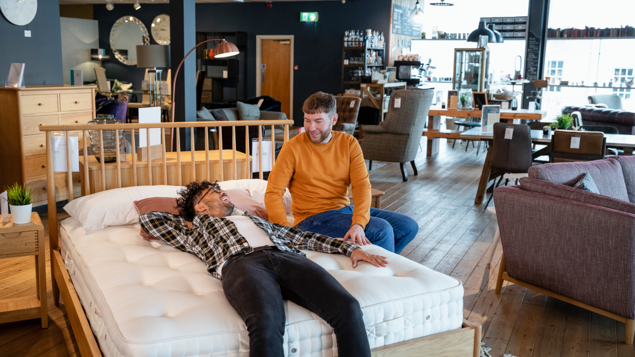 A mid adult couple viewing a home furniture showroom together, one man is lying on a bed while his fiancé sits near him on the bed. They are testing out the bed and considering buying it.
