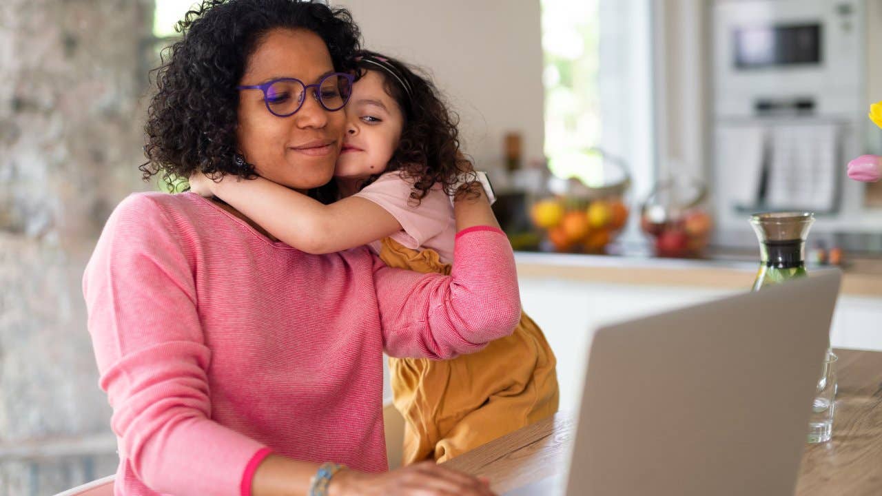 Multiracial girl with her mother hugging, having fun in kitchen during home-office and homeschooling time.