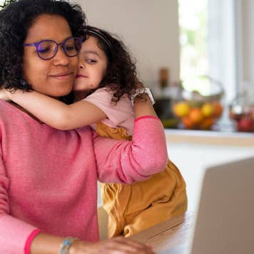 Multiracial girl with her mother hugging, having fun in kitchen during home-office and homeschooling time.