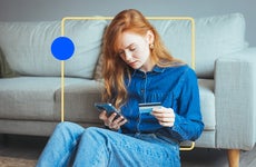 design element of an image of a woman sitting in front of a couch holding a phone in one hand and credit card in the other