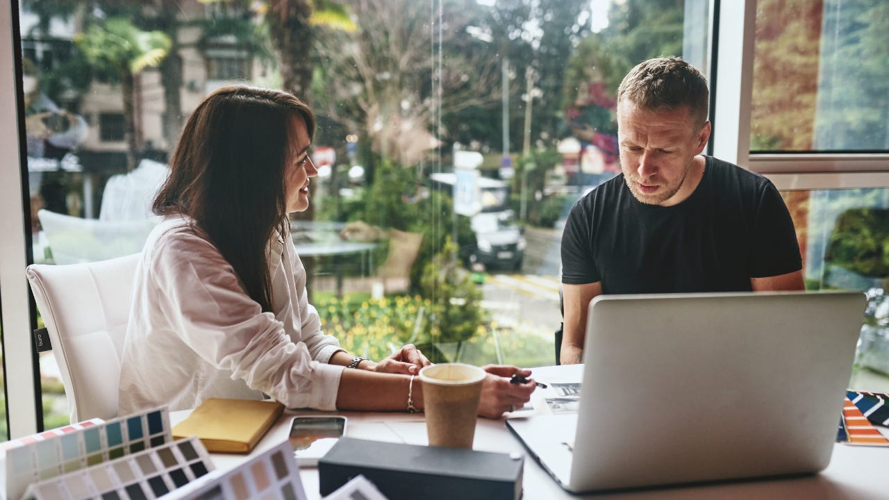 A business owner sits in her office and patiently explains something to her partner, who sits looking slightly confused.