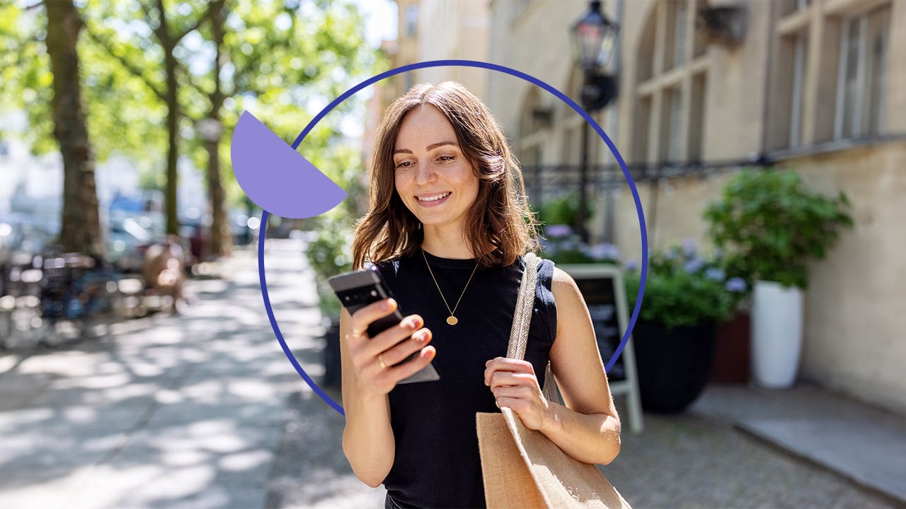 Young woman smiling and walking down a city street with here cellphone out