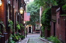 The narrow cobblestoned Acorn Street with Federal-style red row houses, with brick sidewalks and lit by gas lanterns at dawn, with an American flag in the historical neighbourhood of Beacon Hill
