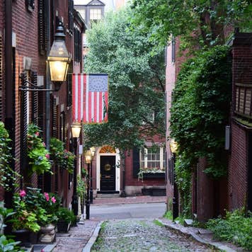 The narrow cobblestoned Acorn Street with Federal-style red row houses, with brick sidewalks and lit by gas lanterns at dawn, with an American flag in the historical neighbourhood of Beacon Hill