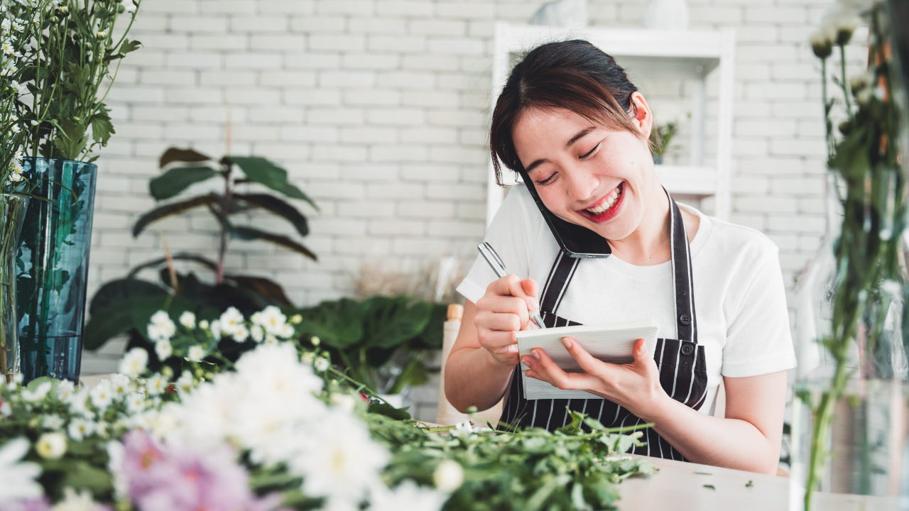 A smiling florist in a striped apron listens on the phone while taking notes in a notepad.