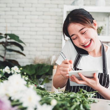 A smiling florist in a striped apron listens on the phone while taking notes in a notepad.