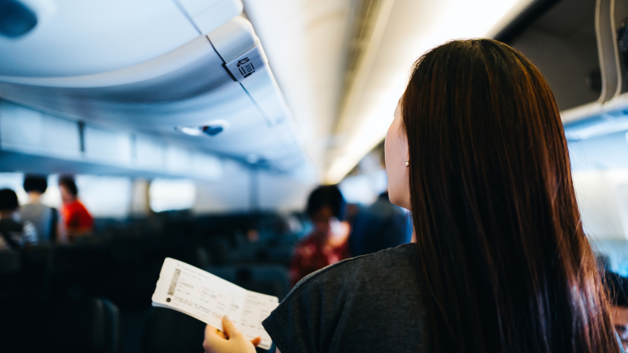 Rear view of young Asian female traveller holding her boarding pass, walking down the aisle in the airplane looking for her seat. Travel and vacation concept