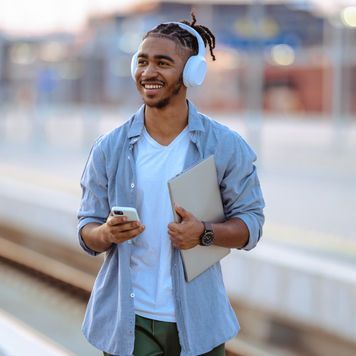 black man with headphones and holding a laptop, smiling with a phone in his other hand