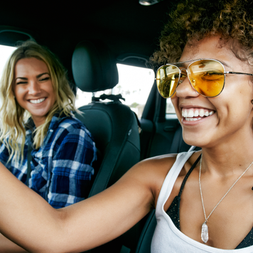 Portrait of two women with long blond and brown curly hair sitting in car, wearing sunglasses, smiling.