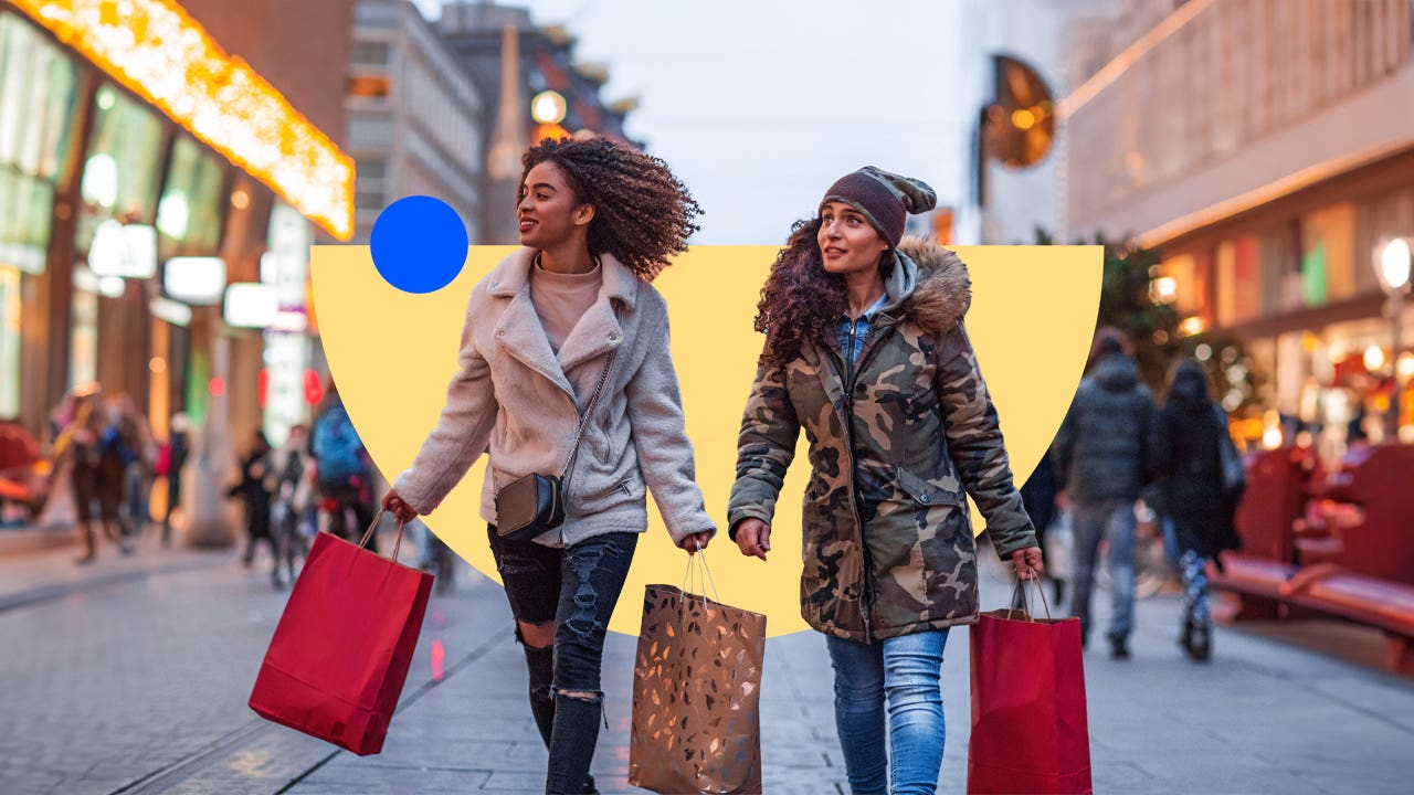 Two young holiday shoppers holding shopping bags walk outside along a shopping plaza.