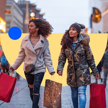 Two young holiday shoppers holding shopping bags walk outside along a shopping plaza.