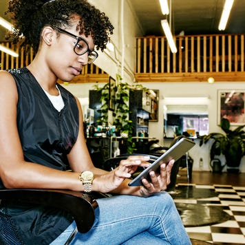 A young woman sits in the chair at her empty salon, looking at her tablet.