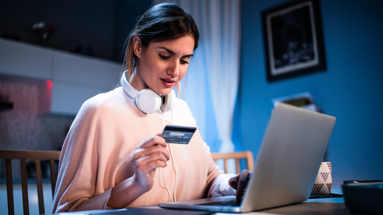 women sitting down in front of her laptop with headphones around her neck holding a credit card in her hand