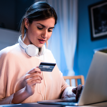 women sitting down in front of her laptop with headphones around her neck holding a credit card in her hand