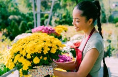A young, smiling entrepreneur is outdoors at her garden center, holding a container full of potted flowers.