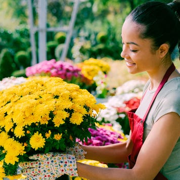 A young, smiling entrepreneur is outdoors at her garden center, holding a container full of potted flowers.