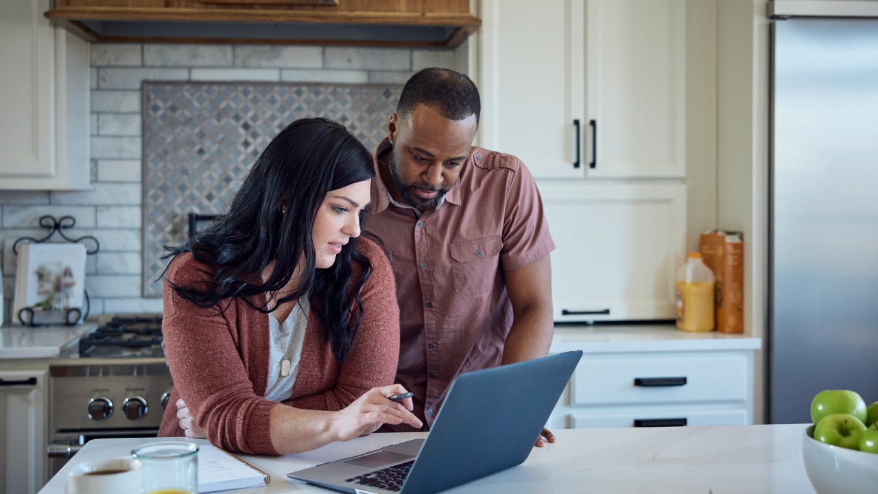 A couple stand close together at the counter in their kitchen, looking at a laptop.