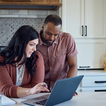 A couple stand close together at the counter in their kitchen, looking at a laptop.