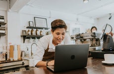 A smiling coffee shop owner leans over the counter, working on a laptop.
