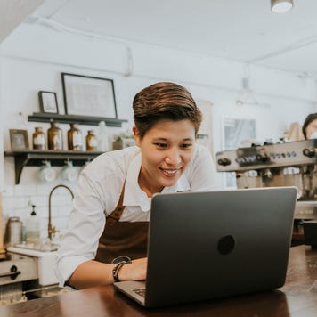 A smiling coffee shop owner leans over the counter, working on a laptop.