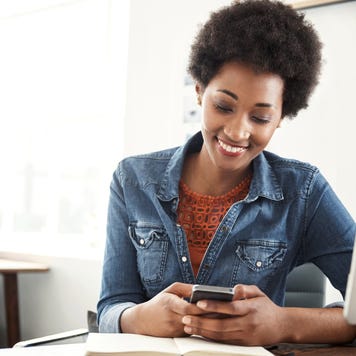 A smiling woman sits at her desk looking at her cellphone.