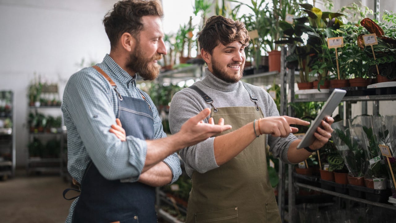Two men in aprons look at a tablet inside a garden center.