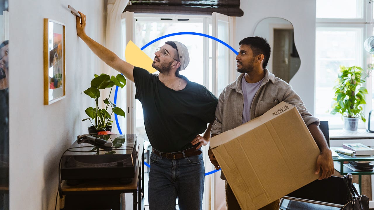 Two men standing together inside a house. One is holding a box.