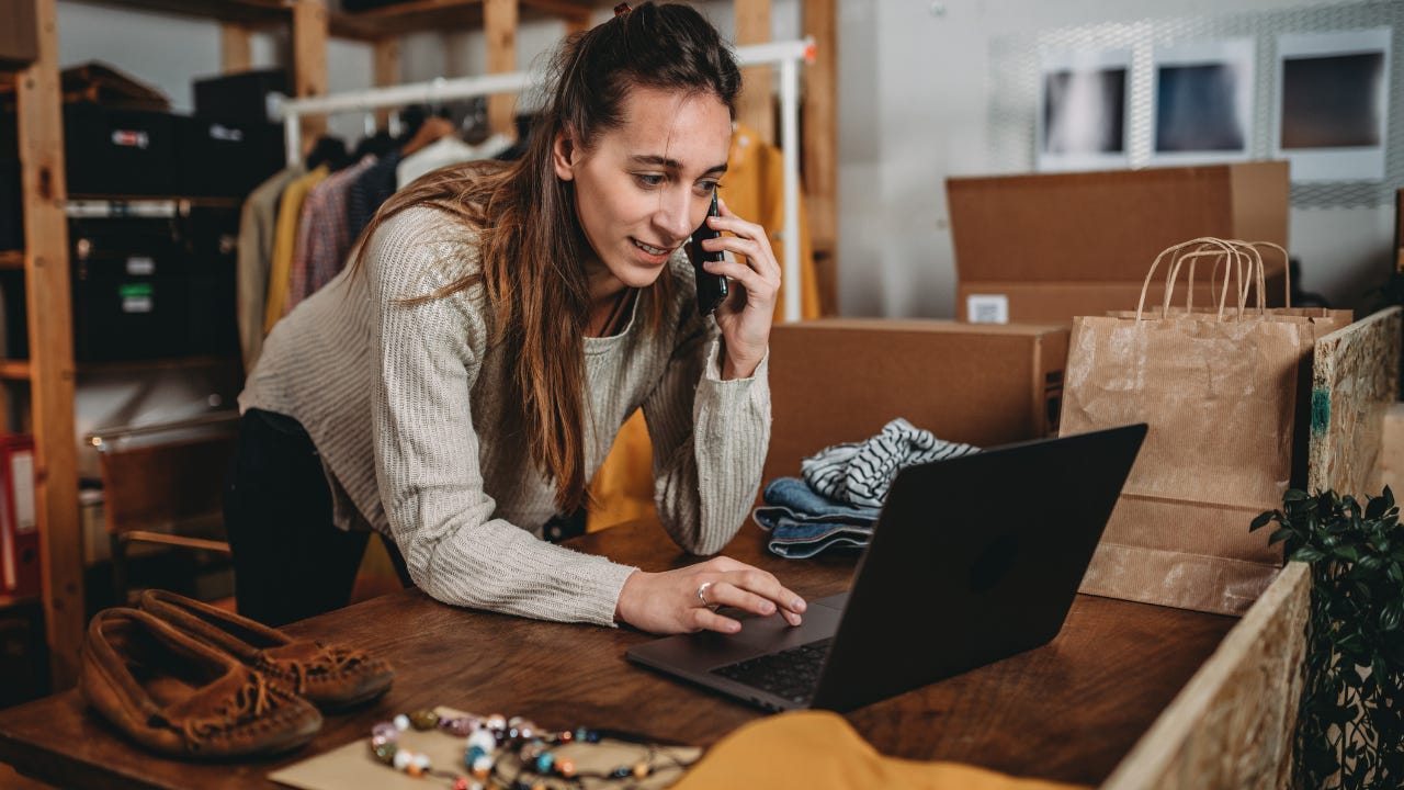 A woman leans over a table in a shop, looking at her laptop while talking on the phone.