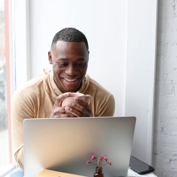 Man sitting at cafe table holding coffee cup and looking at laptop, smiling
