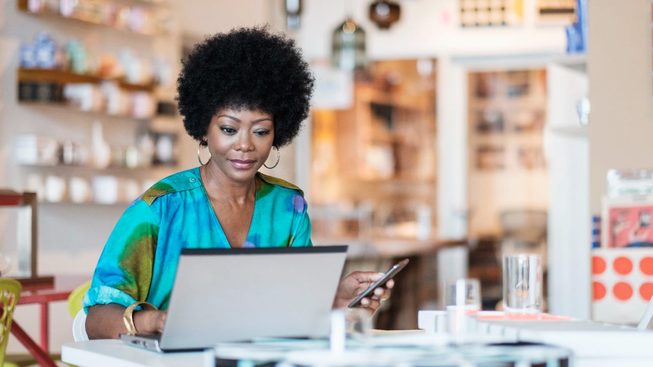 Black woman business owner sits in front of a laptop holding a cell phone.