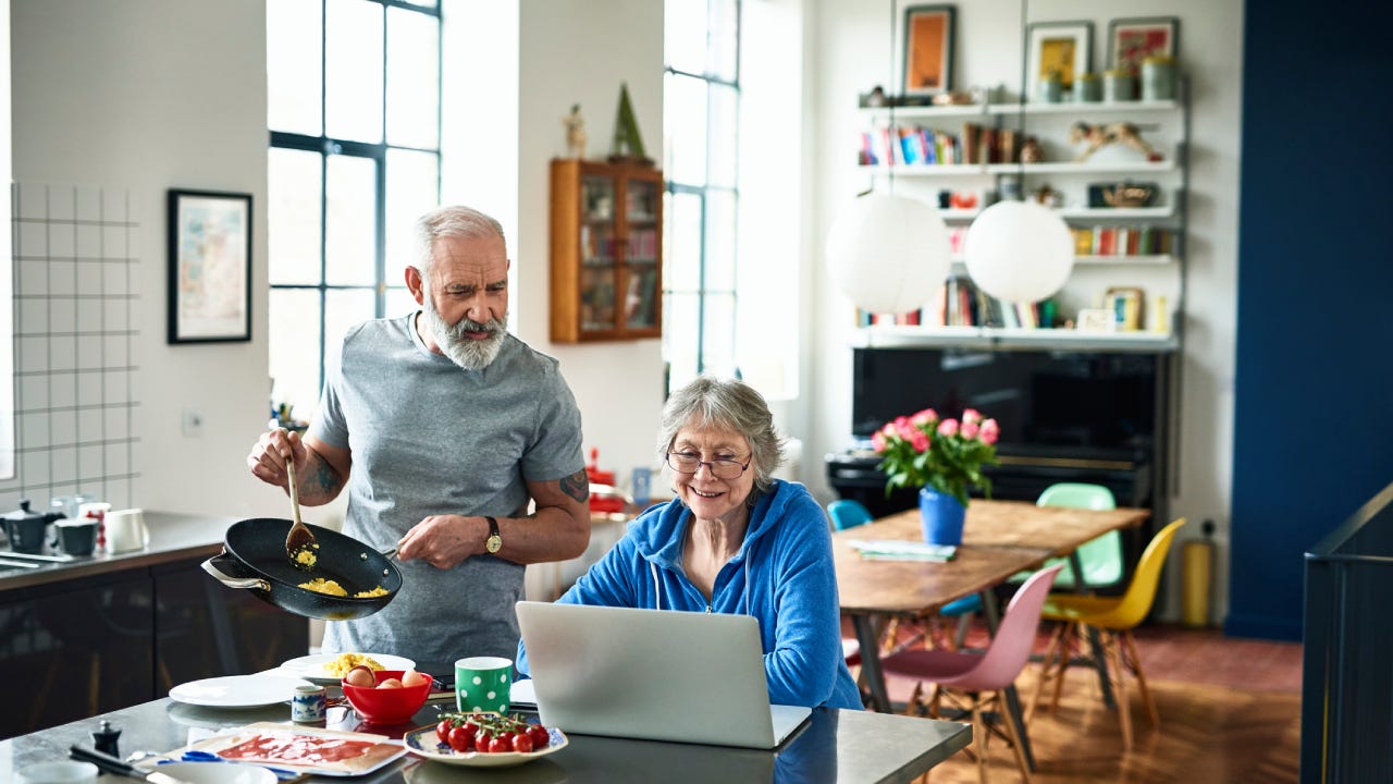 Senior woman using laptop and smiling as man serves breakfast